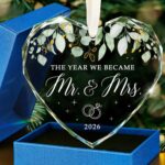 HEXMOZ Mr & Mrs 2026 heart-shaped glass ornament in a navy blue gift box with an ivory ribbon, hanging.