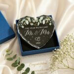 HEXMOZ Mr & Mrs 2026 heart-shaped glass ornament in a navy blue gift box with an ivory ribbon, hanging.