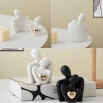 A white ceramic abstract statue depicting two figures hugging, with a golden heart detail, placed on a surface.