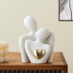 A white ceramic abstract statue depicting two figures hugging, with a golden heart detail, placed on a surface.