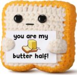 Close-up of a small, square, light brown crochet toast plush with embroidered eyes and a red heart, holding a white banner th