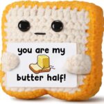 Close-up of a small, square, light brown crochet toast plush with embroidered eyes and a red heart, holding a white banner th