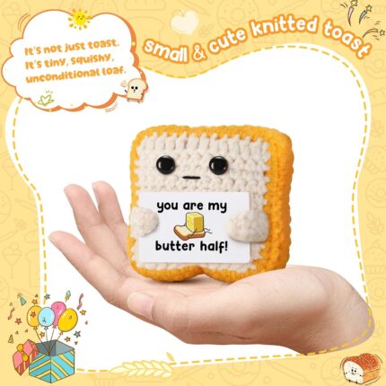 Close-up of a small, square, light brown crochet toast plush with embroidered eyes and a red heart, holding a white banner th
