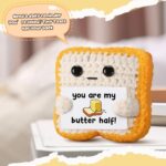 Close-up of a small, square, light brown crochet toast plush with embroidered eyes and a red heart, holding a white banner th