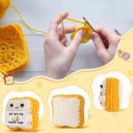 Close-up of a small, square, light brown crochet toast plush with embroidered eyes and a red heart, holding a white banner th